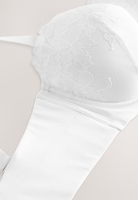Soutien-gorge en dentelle blanche avec motif en maille florale sur les bonnets et bande en tissu lisse sur fond neutre.