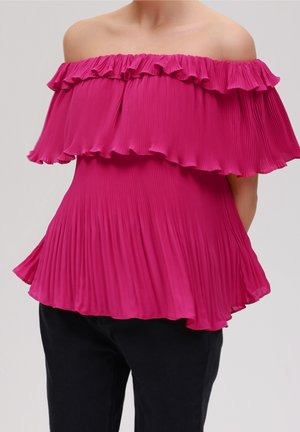 Haut plissé rose vif à épaules dénudées avec volants superposés, porté avec un pantalon noir sur une personne devant un fond uni.