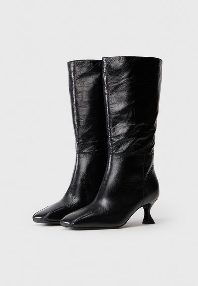 Páros, középmagas fekete bőr csizmák alacsony, ívelt sarkakkal és hegyes orral, sima világos háttér előtt bemutatva.