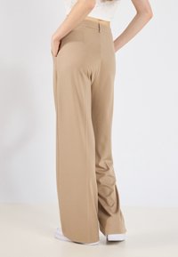 Beige wijde broek gemaakt van een gladde stof, met zijzakken en een platte voorkant. Ze worden gedragen met witte schoenen.