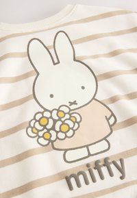 Tissu rayé blanc et beige avec un lapin dessin animé tenant un bouquet de fleurs blanches et jaunes et le mot « miffy » en dessous.