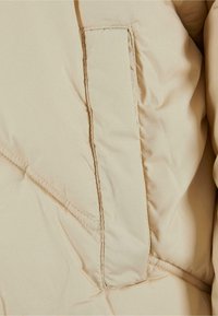 Beige puffer jacket close-up, featuring a stitched pocket, smooth texture, and diagonal stitching pattern along the fabric.