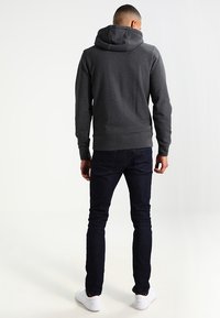 Haut à capuche gris foncé avec un cordon de serrage, des poignets côtelés et un ourlet ; associé à un jean en denim foncé et des baskets blanches. Détails de couture visibles.