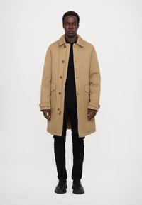 Beige long coat with a button-front design, side pockets, and a collar. Worn over a black top and black pants, complemented by dark shoes.