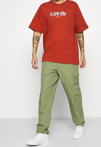 T-shirt en coton rouge avec un graphique coloré "Levi's". Associé à un pantalon cargo vert avec poches latérales et des baskets blanches.