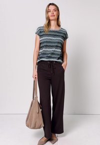 Femme debout portant un haut à manches courtes avec un motif zigzag, un pantalon noir, tenant un grand sac fourre-tout beige et portant des ballerines beiges.