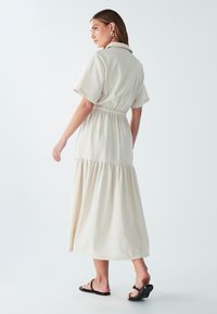 Bežinė midi suknelė su apykakle, trumpomis rankovėmis, elastinga juosmeniu ir sluoksniuotu sijonu. Pagaminta iš lengvos medžiagos su lygiu paviršiumi.