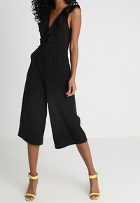 Svart, ärmlös jumpsuit med djup V-ringning och volangdetaljer; culotte-stil på benen; matchad med gula klacksandaler.