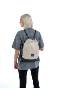 Beige és fekete szövetből készült húzózsinóros hátizsák, állítható pántokkal, minimalista dizájnnal és elől egy márkacímkével.