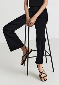 Personne portant un pantalon noir court et des sandales noires à lanières avec des accents dorés, assise sur un haut tabouret noir contre un fond blanc.