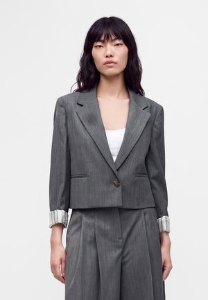 Frau mit langen dunklen Haaren, die einen grauen kurzen Blazer und passende hoch taillierte, weit geschnittene Hosen über einem weißen Oberteil trägt.