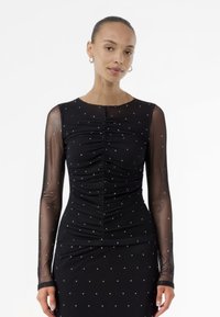 Μαύρο φόρεμα με μακριά διαφανή μανίκια, πτυχώσεις μπροστά, διακοσμημένο με μικρές ασημένιες κουκκίδες σε όλη του την επιφάνεια. Στενή σιλουέτα; στρογγυλή λαιμόκοψη.