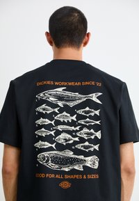 Black cotton t-shirt with white fish illustrations on the back, orange text above stating "DICKIES WORKWEAR SINCE '22" and below, "GOOD FOR ALL SHAPES & SIZES."