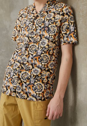 Person trägt ein schwarzes Kurzarmshirt mit gelbem und weißem Blumen- und Pilzmuster und senfgelben Hosen, lehnt an einer Wand.