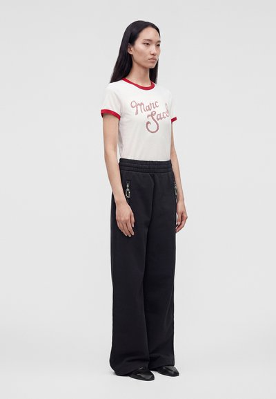 Γυναίκα όρθια, φοράει λευκό μπλουζάκι Marc Jacobs με κόκκινο τελείωμα, μαύρο φαρδύ παντελόνι με λεπτομέρειες φερμουάρ και μαύρα παπούτσια, μπροστά σε απλό φόντο.