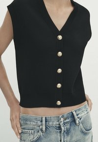 Mustat neulotut hihattomat topit, joissa on V-pääntie, kuusi kultanappia edessä ja lyhyt malli. Yhdistetty vaaleneviin farkkushortseihin.