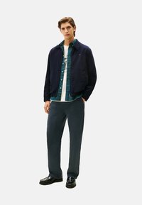 Marineblauwe fluwelen jas over een groen geruit shirt, witte T-shirt en donkere broek, gedragen met zwarte schoenen. Eenvoudige, gelaagde look.