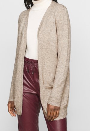 Femme portant un cardigan beige ouvert sur un col roulé blanc et un pantalon en cuir bordeaux à cordon, debout devant un fond uni.