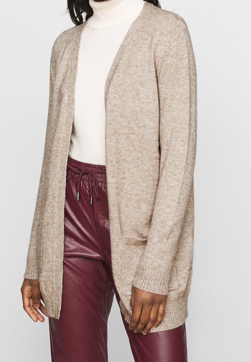 Femme portant un cardigan beige ouvert sur un col roulé blanc et un pantalon en cuir bordeaux à cordon, debout devant un fond uni.