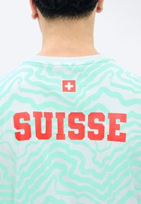 Henkilön takanäkymä, jossa on vaaleansininen paita, jossa on vedenvihreitä aaltoilevia kuvioita ja punainen teksti "SUISSE" punaisen neliön alla, jossa on valkoinen risti.