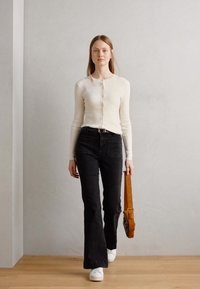 Bela cardigan s gumbi, črni široki jeans in bele superge. Rjava torba za ramo z naramnicami, lesena tla in siva stena v ozadju.