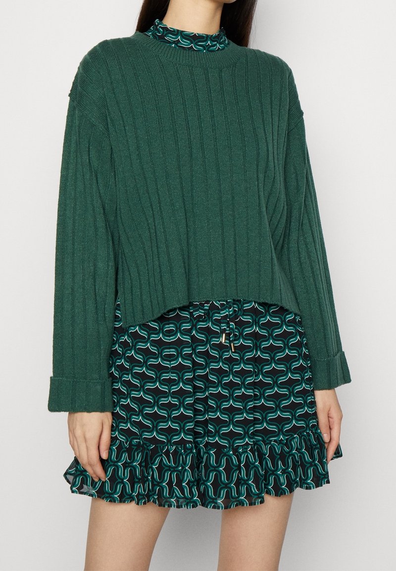 Maglione verde a costine con collo alto, indossato sopra una gonna nera con un motivo astratto verde acqua e orlo arricciato.