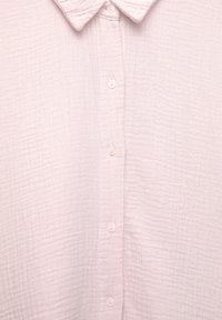 Chemise en coton texturé rose clair avec col et six boutons devant, montrant les détails du tissu et les plis doux.