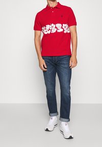 T-shirt polo rouge avec un motif floral blanc à la taille, manches courtes et col. Associé à un jean bleu et des baskets blanches.