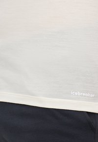 Lys beige kortermet skjorte laget av mykt materiale, med en hvit "icebreaker"-logo nær kanten, kombinert med mørk stoff under.