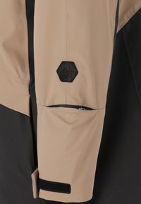 Beige och svart jacka med en hexagonal logotyp, en dragkedjeficka på ärmen, och en texturerad tygfinish.