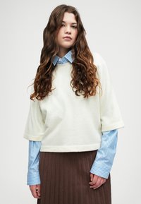 White, textured short-sleeve sweater layered over a blue and white striped long-sleeve shirt, paired with a brown pleated skirt.