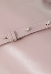 Primo piano di una borsa in vera pelle rosa con una texture liscia, una tracolla con due borchie simili a perle e una leggera lucentezza.