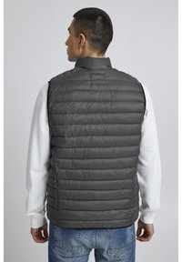 Chaleco de plumas gris con cuello alto, diseño acolchado y sin mangas, usado sobre una camiseta de manga larga blanca. Textura suave y ligero acolchado.