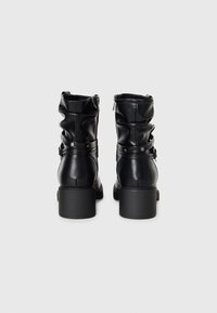 Paire de bottes en cuir noires mi-mollet à talons carrés, zips latéraux et brides décoratives avec clous métalliques sur fond clair.