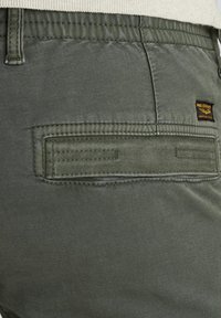 Olivgrüne Hose mit glatter Textur, die eine Gesäßtasche mit horizontaler Naht und einem kleinen Markenlabel in Schwarz und Gold aufweist.