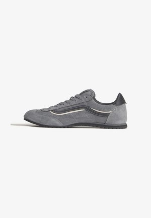 Sneaker grigia in camoscio con lacci, banda laterale bianca e nera e suola in gomma nera, vista di profilo su sfondo bianco.