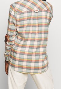 Person wearing a pastel plaid long-sleeve shirt and off-white pants, standing with one arm bent behind the back.