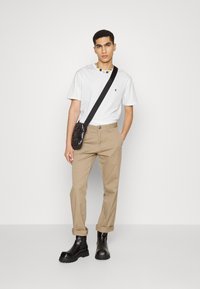 Witte T-shirt met korte mouwen en een klein logo, beige broek en zwarte enkellaarzen met rubberen zolen. Schoudertas van glanzend zwart materiaal.
