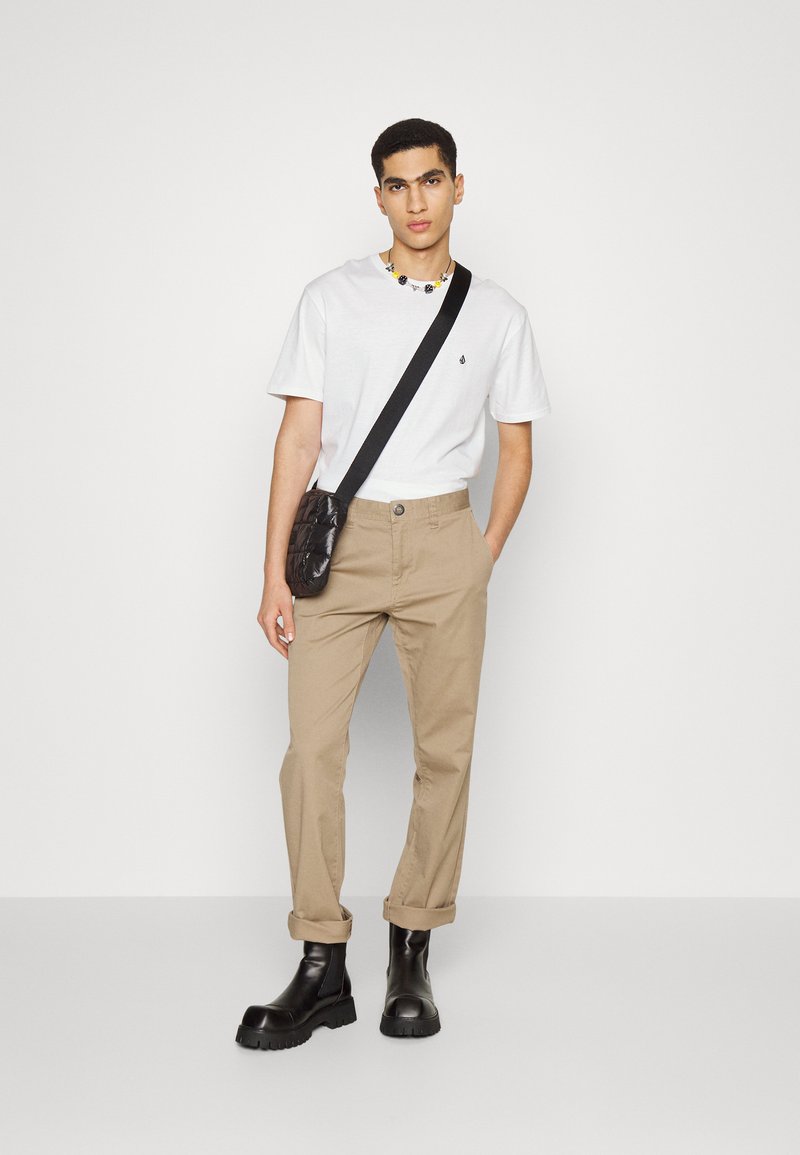 Weißes T-Shirt mit kurzen Ärmeln und kleinem Logo, beigem Hosen, und schwarzen Gummisohlen-Stiefeletten. Umhängetasche aus glänzendem schwarzem Material.