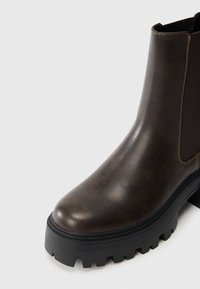 Rudos odinės kulkšnės botai su apvaliu pirštu, elastingu šoniniu panele ir storu juodu guminiu padu, kuriame yra rašto motyvas. Lygi tekstūra su siuviniais.