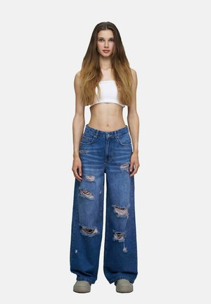 Jeune femme aux longs cheveux portant un crop top blanc, un jean bleu déchiré et ample, et des chaussures beiges, debout devant un fond uni.