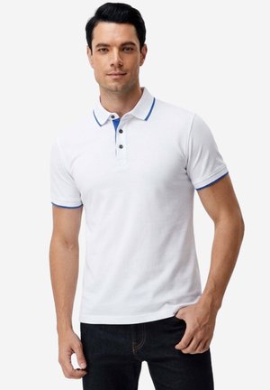 Uomo che indossa una polo bianca a maniche corte con bordatura blu sul colletto e sulle maniche, abbinata a jeans scuri, in piedi con una mano in tasca.