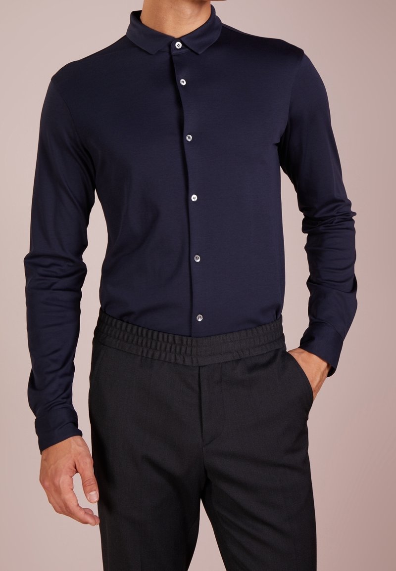 Homme portant une chemise bleu marine ajustée à manches longues boutonnée, rentrée dans un pantalon noir à taille élastique, une main dans la poche.