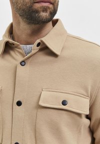 Skäggig man iförd en beige knappad skjortjacka med svarta tryckknappar över en grå stickad tröja, visad från axlarna till läpparna.