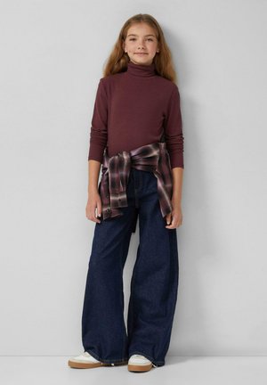 Mädchen mit langen Haaren, das einen weinroten Rollkragenpullover, weit geschnittene dunkle Jeans, weiße Sneakers und ein karierter Hemd um die Taille gebunden trägt.