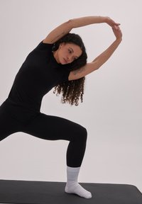 Zwart aansluitend topje en leggings met ribbeltextuur. Witte enkelsokken. Het model bevindt zich in een zijwaartse rekpositie op een zwart yogamat.