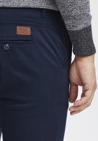 Pantalones azul marino de algodón con una textura suave, que cuentan con un parche de cuero marrón y un cierre de botón en el bolsillo delantero.