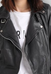 Veste de motard en cuir noir avec quincaillerie en argent, fermeture éclair asymétrique et larges revers, superposée sur un t-shirt graphique blanc.