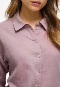 Světle růžová, strukturovaná košile na knoflíky s límcem a šesti shodnými knoflíky vpředu, se viditelným mačkaným vzorem.