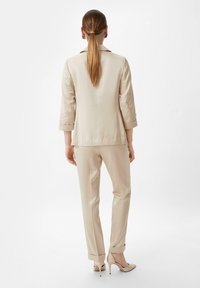 Beige tailored jacket with rolled sleeves, paired with matching tailored pants. Smooth fabric, slim fit, and subtle sheen. Elegant high heels.